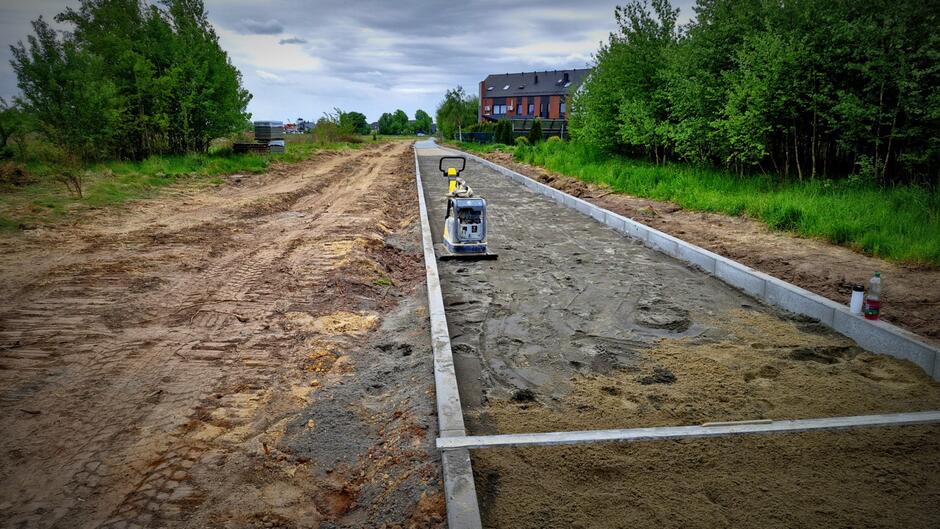 na zdjęciu gruntowa droga, po prawej widać układane krawężniki, w tle zielone drzewa