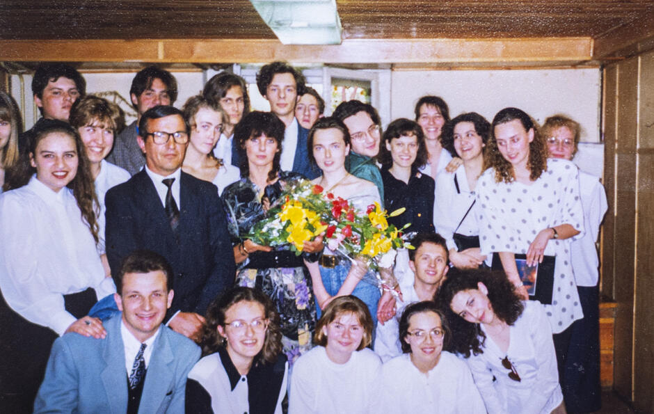 To kolorowe zdjęcie grupowe z lat 90. XX wieku, wykonane wewnątrz budynku – najpewniej w jednej z sal III Liceum Ogólnokształcącego w Gdańsku – ukazuje klasę maturalną z nauczycielami w wyjątkowym momencie, prawdopodobnie podczas zakończenia roku szkolnego lub pożegnania nauczycielki.
