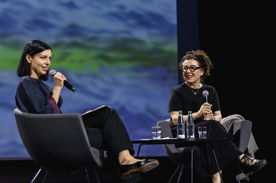 Dwie kobiety siedzą na fotelach na scenie i rozmawiają.