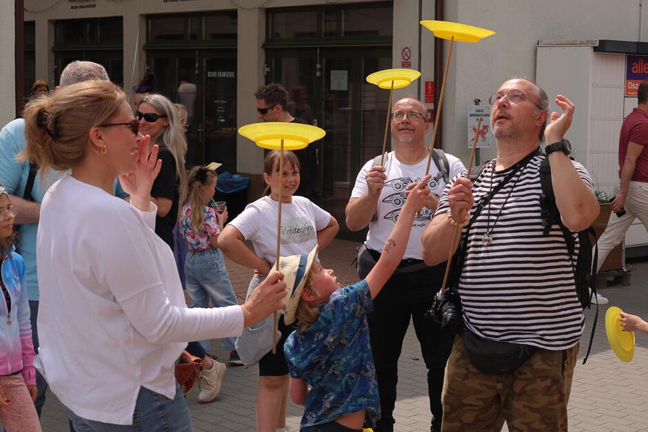 Na chodniku stoi kilka osób, dorośli i dzieci i kręcą żółtymi talerzykami na patykach podczas festynu.