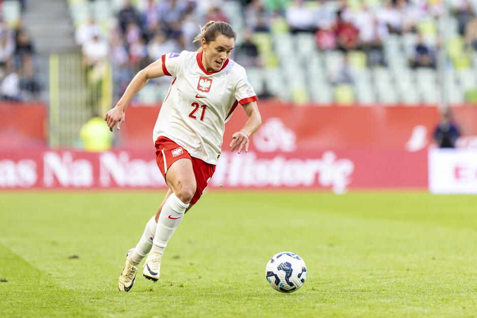 młoda kobieta w dwu częściowym stroju sportowym biegnie po murawie kopiąc piłkę za nią widoczne trybuny z kibicami