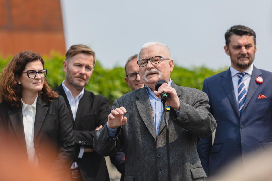 na zdjęciu lech wałęsa w marynarce, mówi do mikrofonu, za nim stoi kilka osób