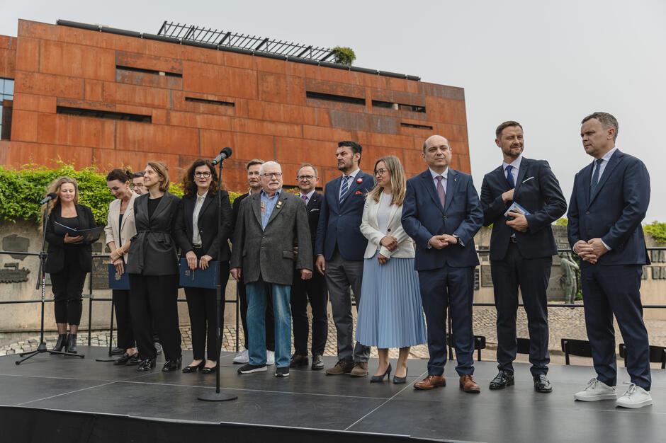 na zdjęciu scena na której stoi kilkanaście dorosłych osób w oficjalnych strojach