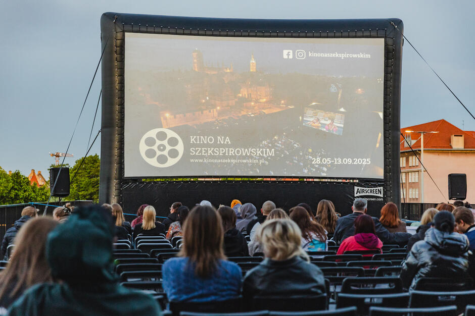 Ludzie siedzą na zewnątrz, oglądając film na dużym ekranie kinowym w ramach plenerowego wydarzenia „Kino na Szekspirowskim”. Na ekranie widać zapowiedź cyklu seansów, który trwa od 28 maja do 13 września 2025 roku