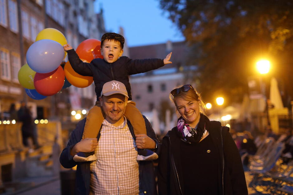 Kobieta iu mężczyzna z dzieckiem na barana pozują z balonami do zdjęcia, stojąc na ulicy starego miasta. 