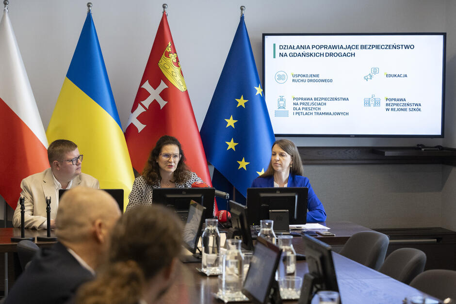 Na zdjęciu widzimy konferencję prasową poświęconą poprawie bezpieczeństwa na drogach w Gdańsku. Prezentacja na ekranie za uczestnikami zawiera główne obszary działań: Uspokojenie ruchu drogowego Poprawa bezpieczeństwa na przejściach dla pieszych i pętlach tramwajowych Poprawa bezpieczeństwa w rejonie szkół Edukacja