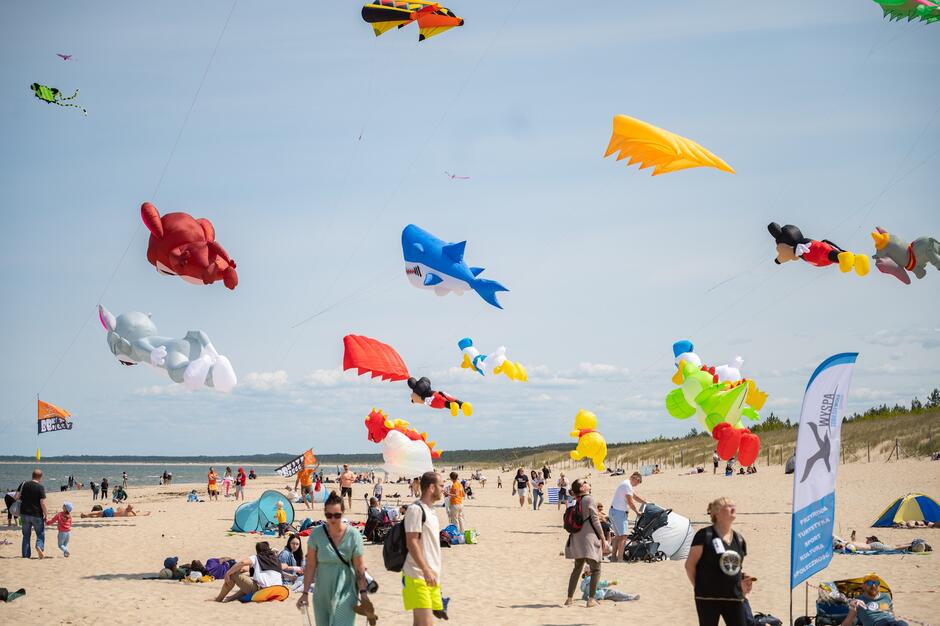 Na zdjęciu widać plażę pełną ludzi, nad którą unoszą się kolorowe, ogromne latawce w kształcie postaci z kreskówek i zwierząt. Panuje radosna, rodzinna atmosfera, a błękitne niebo i słoneczna pogoda sprzyjają zabawie podczas festiwalu latawców
