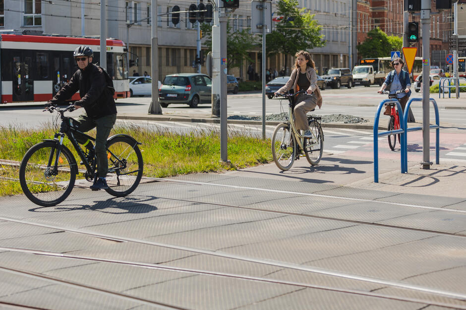 Zdjęcie ukazuje trzech rowerzystów przejeżdżających przez skrzyżowanie w miejskim otoczeniu, z widocznym torowiskiem tramwajowym i sygnalizacją świetlną dla rowerów. Scena oddaje codzienny charakter ruchu rowerowego w mieście i promuje bezpieczne poruszanie się po dobrze zorganizowanej infrastrukturze rowerowej.