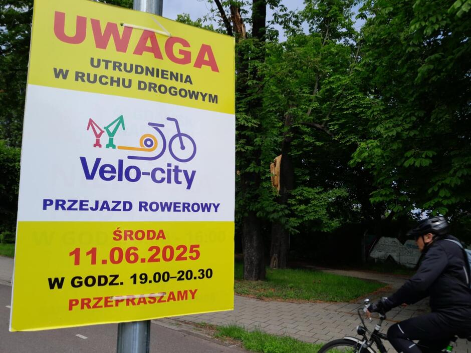 Zdjęcie przedstawia informacyjny plakat ostrzegający o utrudnieniach w ruchu drogowym z powodu rowerowego przejazdu „Velo-city”, który odbędzie się w środę 11 czerwca 2025 roku w godzinach 19:00–20:30. Obok plakatu, na tle zielonych drzew, jedzie rowerzysta ubrany w czarny strój i kask, co podkreśla aktywny charakter wydarzenia.