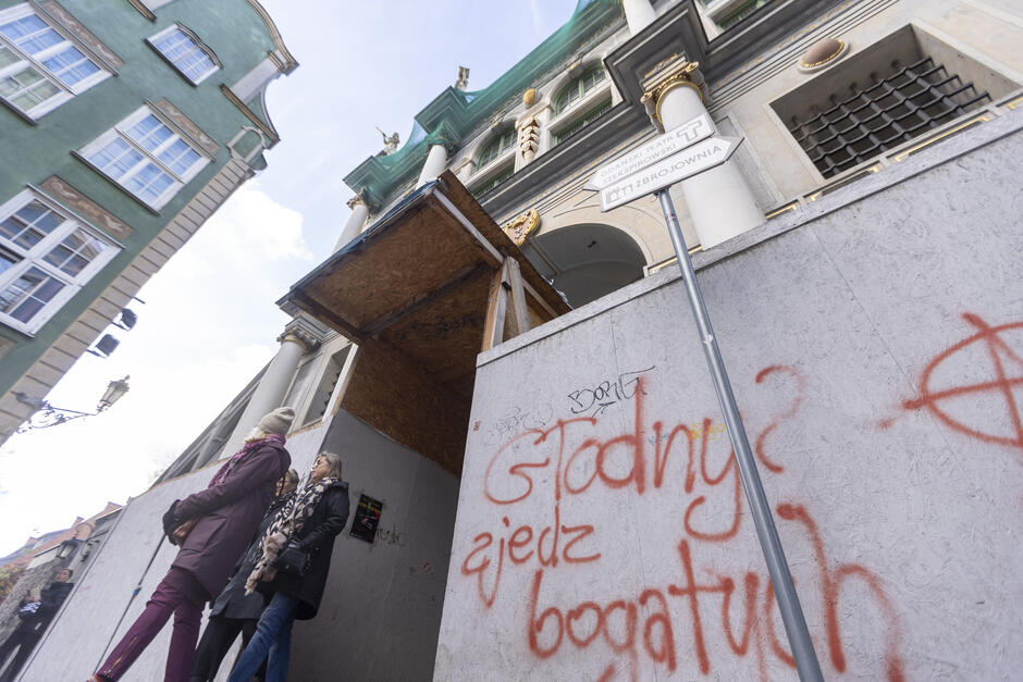 Na zdjęciu widoczna jest Złota Brama w Gdańsku — jeden z najbardziej znanych zabytków miasta, pochodzący z początku XVII wieku. Reprezentuje styl manieryzmu niderlandzkiego, a jego architektem był Abraham van den Blocke. Brama jest obecnie częściowo ogrodzona z powodu prac remontowych — widoczne są białe płyty zabezpieczające teren budowy oraz prowizoryczne zadaszenie przejścia. Niestety, ogrodzenie zostało pokryte wandalicznym graffiti, na którym widnieje m.in. napis  Gdańsk zjedz bogatych , symbol anarchii (litera A w kółku) i skrót  ACAB  (często używany w kontekście sprzeciwu wobec policji).