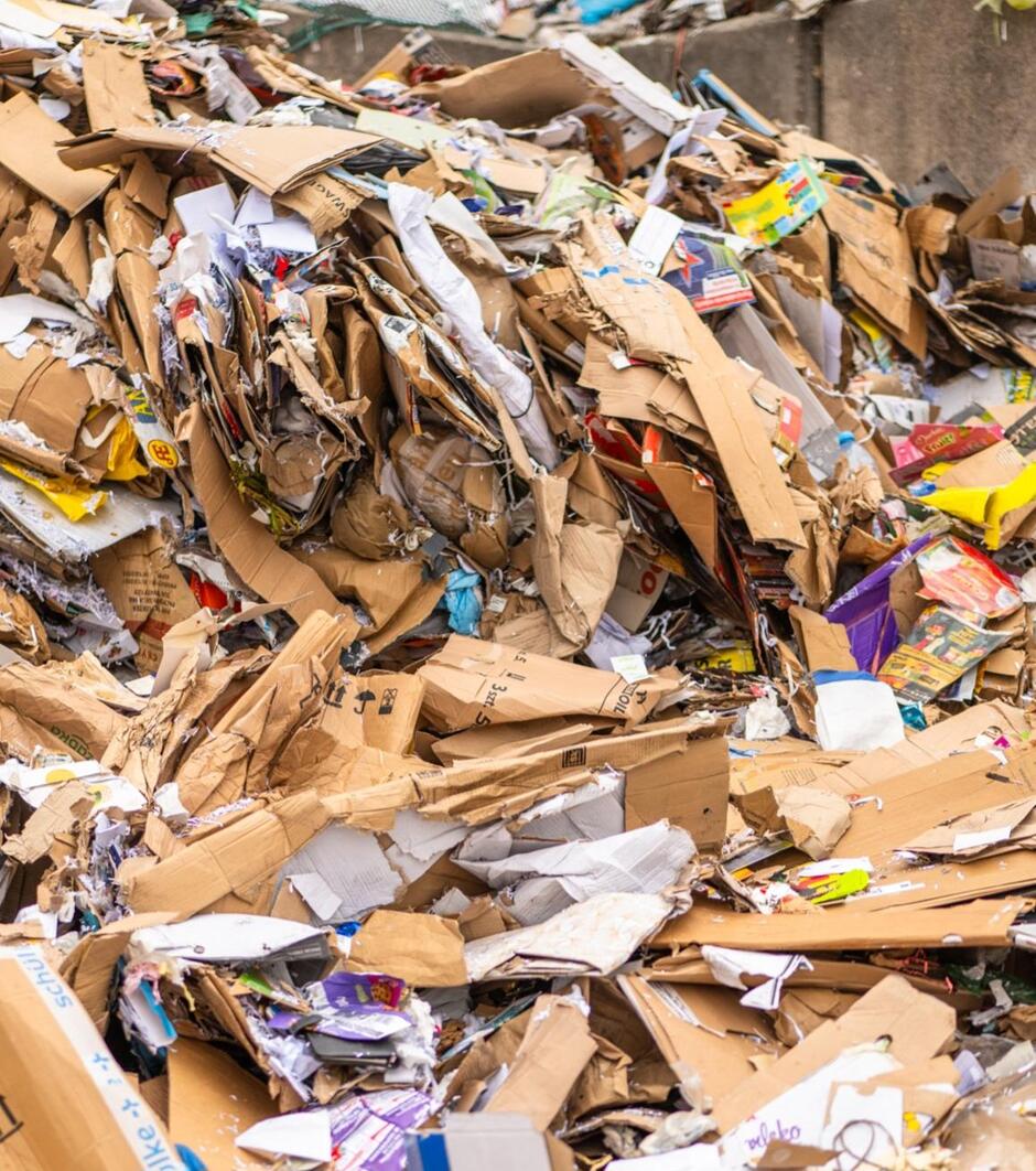 Na zdjęciu znajduje się duża sterta odpadów papierowych, głównie w postaci zużytych kartonów, opakowań i gazet. Widoczna różnorodność materiałów wskazuje na ich pochodzenie z selektywnej zbiórki surowców wtórnych przeznaczonych do recyklingu.