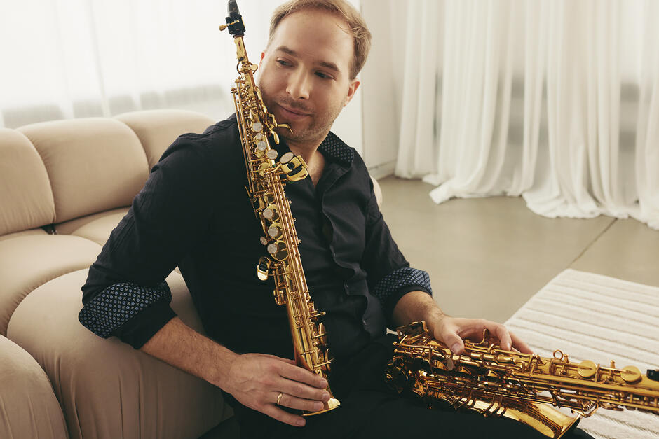 Na zdjęciu mężczyzna w czarnej koszuli, siedzący na kanapie, trzyma w rękach saksofony.