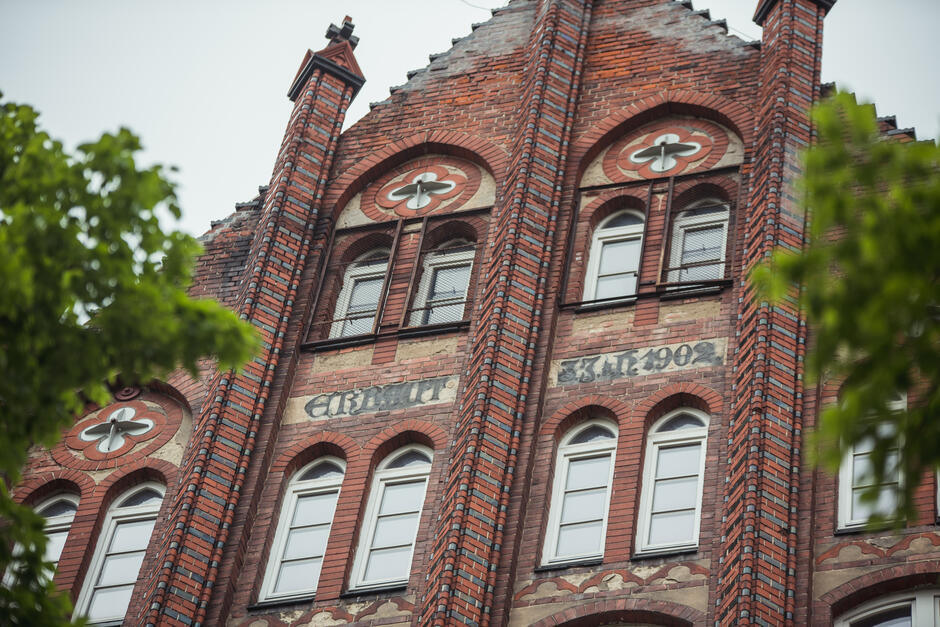 Zdjęcie przedstawia fragment neogotyckiego budynku z czerwonej cegły, ozdobionego charakterystycznymi łukami i detalami z jasnego kamienia oraz cegły w kontrastujących kolorach. Nad środkowymi oknami widnieje napis „Errichtet 33.XI.1902”, wskazujący na datę wzniesienia budynku. 