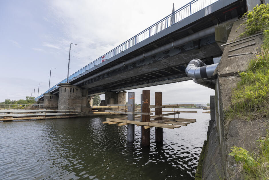 na zdjęciu fragment mostu, pod nim wbitych kilka pali, które wystają z rzeki