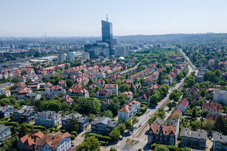 Zdjęcie lotnicze przedstawia dzielnicę Gdańska – Oliwę, z charakterystyczną zabudową willową o czerwonych dachach oraz nowoczesnym kompleksem biurowym Olivia Centre w tle. Widoczna jest harmonia między historyczną architekturą a nowoczesnymi elementami miejskimi, otoczonymi dużą ilością zieleni.