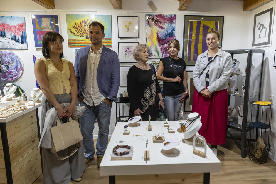 Grupa pięciu osób pozuje wewnątrz galerii sztuki, otoczona kolorowymi obrazami i biżuterią wystawioną na stołach. Wszyscy wyglądają na zadowolonych i gotowych do świętowania otwarcia wystawy