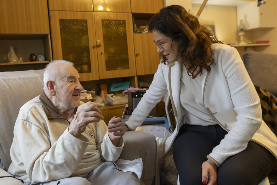 Starszy mężczyzna siedzi w fotelu i rozmawia z uśmiechniętą kobietą, która pochyla się w jego stronę. Atmosfera jest ciepła i pełna wzajemnego zainteresowania, sugerując serdeczne spotkanie rodzinne lub opiekuńcze.