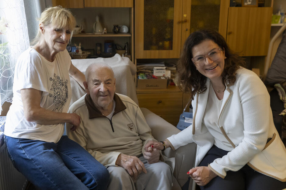 Starszy mężczyzna siedzi w fotelu, trzymając za ręce dwie kobiety – jedna siedzi po lewej, druga kuca po prawej stronie. Wszyscy uśmiechają się do kamery, co nadaje zdjęciu charakter rodzinnej pamiątki lub ważnej wizyty.