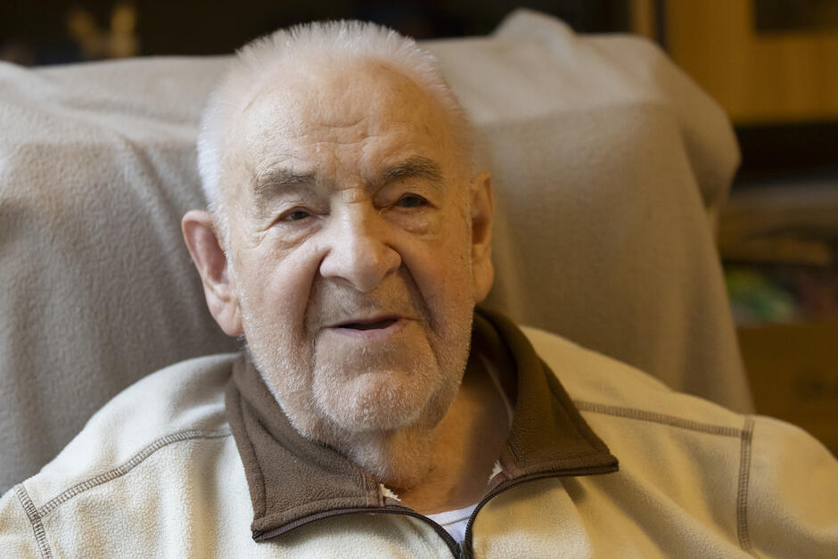 To portret starszego mężczyzny siedzącego w fotelu, z ciepłym i łagodnym wyrazem twarzy. Zdjęcie skupia się na jego twarzy, podkreślając spokój, pogodę ducha i dojrzałość wieku