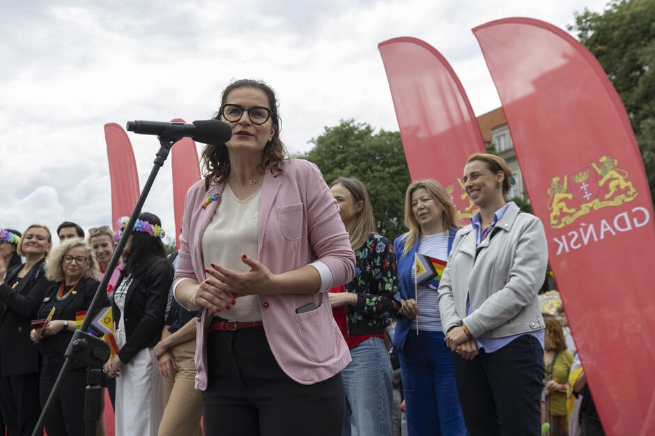 Na tym zdjęciu uchwycony został moment oficjalnego przemówienia podczas Trójmiejskiego Marszu Równości. Kobieta stojąca przy mikrofonie, ubrana w różową marynarkę i białą bluzkę, z tęczową przypinką w klapie, najprawdopodobniej pełni rolę organizatorki, przedstawicielki władz lub zaproszonej gościni. W tle widoczne są czerwone flagi z herbem Gdańska oraz napisem GDAŃSK , co sugeruje, że wydarzenie odbywa się przy wsparciu lub patronacie miasta.