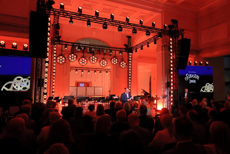 Wnętrze eleganckiej sali koncertowej z czerwonym oświetleniem, scena przygotowana jest do występu muzycznego – widoczna jest orkiestra i publiczność. Na ekranie po prawej stronie znajduje się tytuł "Straszny Dwór", sugerujący, że odbywa się koncert lub spektakl nawiązujący do tej opery Stanisława Moniuszki