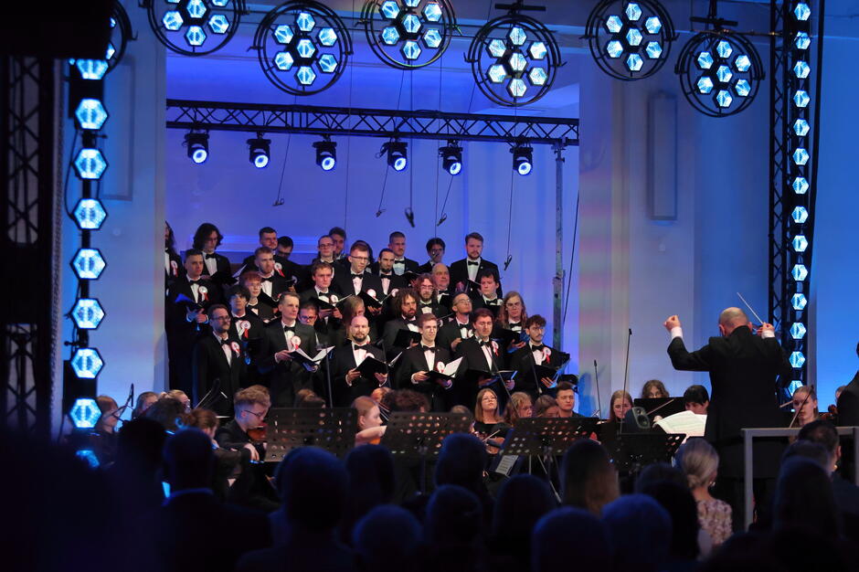 Chór mężczyzn ubranych w smokingi występuje na scenie, z dyrygentem stojącym przodem do nich i orkiestry. Niebieskie oświetlenie i symetryczne rozmieszczenie chórzystów tworzą uroczystą i podniosłą atmosferę koncertu.