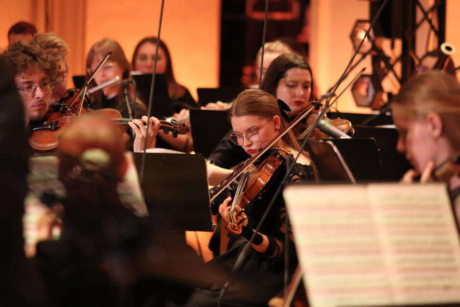 Zbliżenie na muzyków orkiestry, skoncentrowanych na grze – w centrum znajduje się młoda skrzypaczka w okularach, skupiona na nutach. Zdjęcie oddaje intensywność i precyzję pracy muzyków podczas występu.