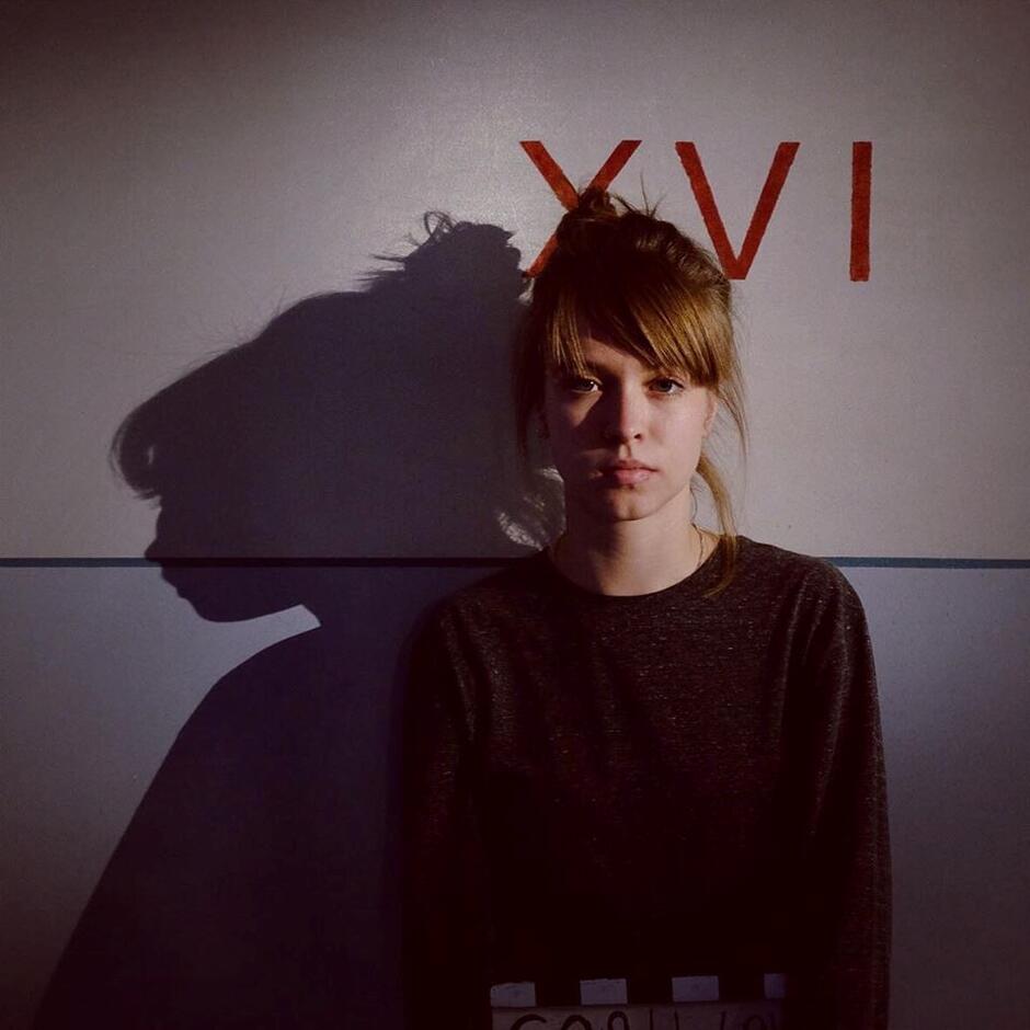 Na zdjęciu kobieta z upiętymi włosami, w tle rzymskie cyfry 16 oraz cień kobiety.