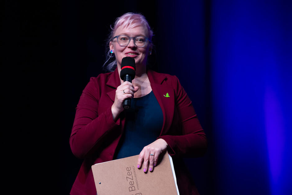 Na zdjęciu kobieta w okularach, w bordowej marynarce, trzymająca w prawej ręce mikrofon, w drugiej teczkę, na tle niebieskiej podświetlonej kurtyny.