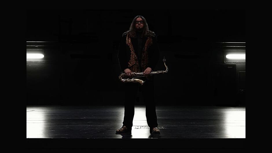Na zdjęciu mężczyzna o długich włosach, w ciemnych okularach, stojący na scenie, trzymający saksofon.