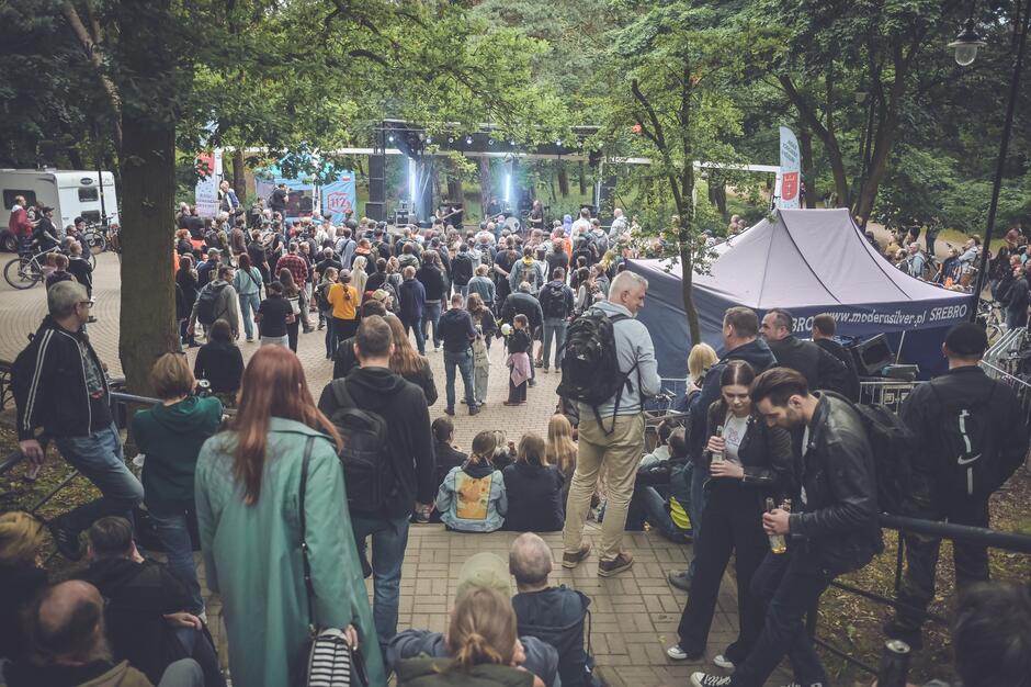 Zdjęcie przedstawia tłum ludzi zgromadzonych na plenerowym koncercie w otoczeniu drzew, prawdopodobnie w ramach festiwalu muzycznego. Na scenie występuje zespół, a w tle widoczne są namioty i banery, m.in. z logo festiwalu Fląder oraz reklamą strony „modernsilver.pl”