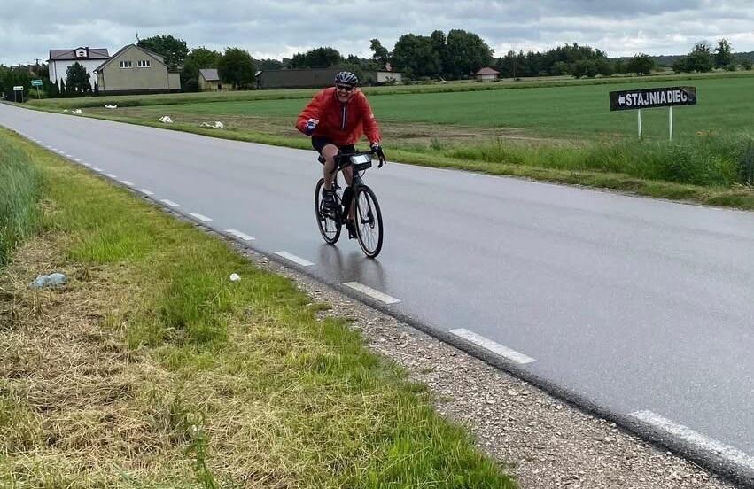 Na zdjęciu widać rowerzystę jadącego pustą, wiejską drogą w otoczeniu zielonych pól i zabudowań. Ubrany w czerwoną kurtkę i kask, uśmiecha się i pozdrawia, co świadczy o pozytywnym nastroju podczas trasy. 