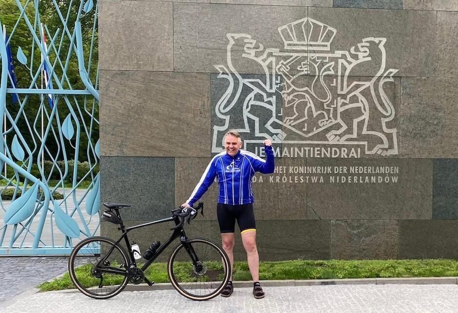 Na zdjęciu widzimy mężczyznę w stroju kolarskim stojącego obok roweru przed budynkiem z herbem Niderlandów i napisem „Je Maintiendrai” oraz „Ambasada Królestwa Niderlandów”. Zadowolony i uśmiechnięty, unosi rękę w geście triumfu, co sugeruje rozpoczęcie lub zakończenie rowerowej wyprawy. 