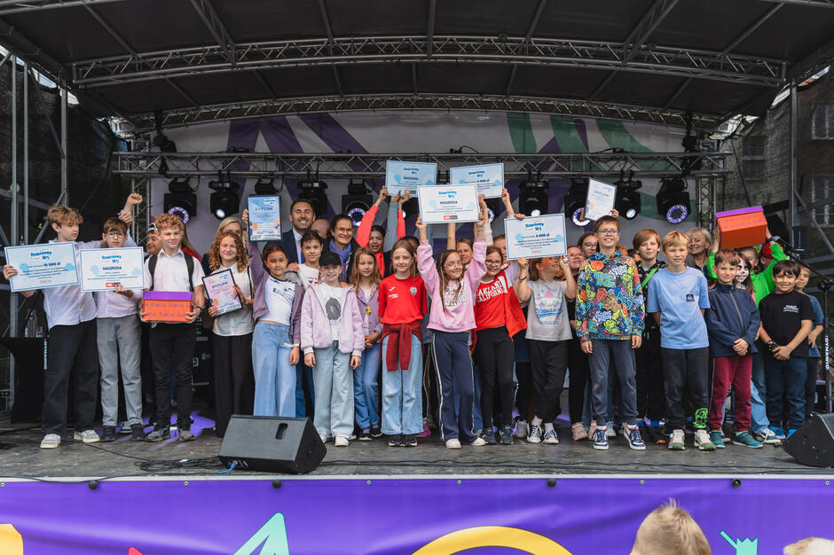Na zdjęciu widać grupę dzieci stojących na scenie, radośnie prezentujących dyplomy, nagrody i symboliczne czeki – prawdopodobnie za udział w konkursie „Rowerowy Maj”. W tle widać dorosłych opiekunów i organizatorów, a cała scena ozdobiona jest kolorowymi dekoracjami, co podkreśla radosny i uroczysty charakter wydarzenia.