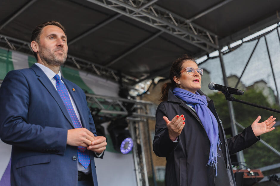Na zdjęciu znajduje się kobieta przemawiająca do mikrofonu oraz stojący obok niej mężczyzna w garniturze – oboje wyglądają na organizatorów lub gości honorowych wydarzenia. Ich poważne, a zarazem serdeczne wyrazy twarzy sugerują, że biorą udział w oficjalnej części uroczystości, najpewniej związanej z wręczeniem nagród lub podsumowaniem inicjatywy.