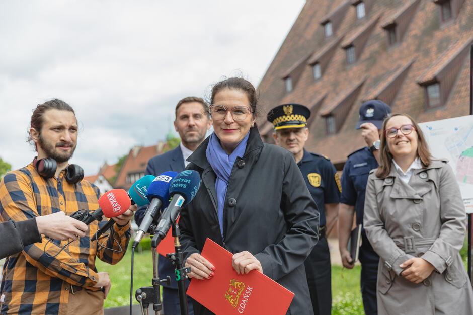 na zdjęciu kobieta w średnim wieku, mówi do mikrofonów, trzyma czerwoną podkładkę w ręce, za nią stoi kilka osób