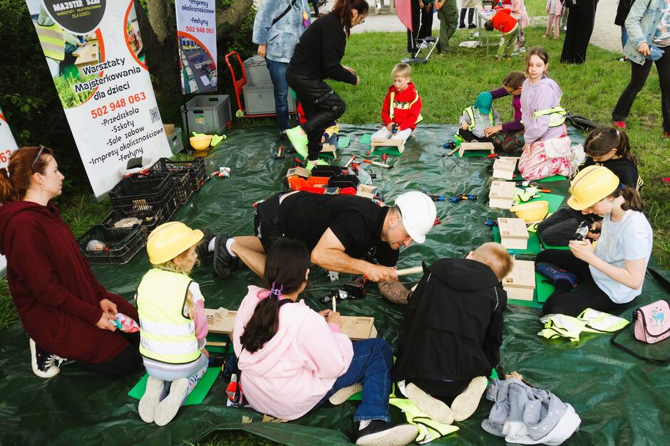 Mężczyzna w kasku i z młotkiem pokazuje zebranym w na macie wokół niego dzieciom jak robić zabawkę