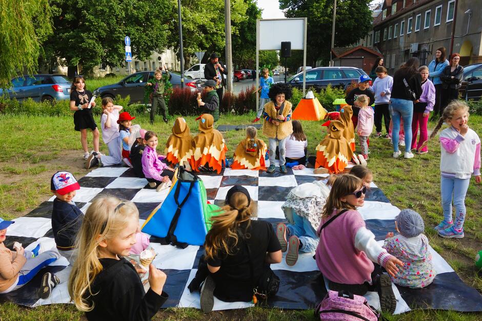 Grupa dzieci w przebraniach siedzących na wielkiej macie w szachownicę