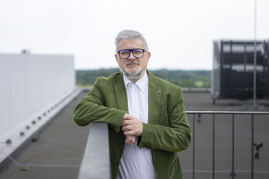 Na zdjęciu widzimy mężczyznę w średnim wieku stojącego na dachu budynku lub tarasie z widokiem na otwartą przestrzeń w tle. Ma siwe włosy, krótko przystrzyżoną brodę i nosi okulary z ciemnymi oprawkami. Ubrany jest elegancko, lecz z nutą swobody – ma na sobie zieloną marynarkę z aksamitu lub innego miękkiego materiału oraz białą koszulę w drobny wzór. Opiera się o barierkę, patrząc spokojnie w stronę aparatu. W tle widać nowoczesne elementy architektury i zieleń na horyzoncie, co nadaje zdjęciu profesjonalny, ale jednocześnie naturalny charakter.