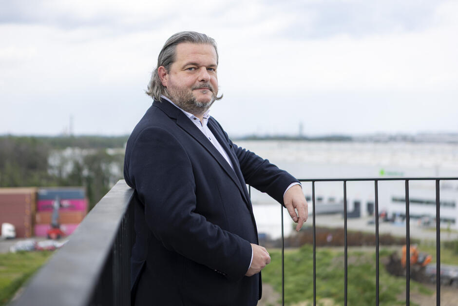 Na drugim zdjęciu widzimy mężczyznę stojącego na tle panoramicznego widoku przemysłowo-logistycznego krajobrazu, prawdopodobnie na tarasie lub dachu budynku. Ma dłuższe, siwiejące włosy, zarost i patrzy spokojnie w stronę obiektywu. Ubrany jest elegancko – ma na sobie ciemnogranatową marynarkę i jasną koszulę bez krawata, co nadaje mu profesjonalny, ale niezbyt formalny wygląd. Opiera się o metalową barierkę, a w tle widoczne są magazyny, tereny zielone i maszyny budowlane, co może sugerować, że jest związany z branżą nieruchomości, przemysłu lub inwestycji infrastrukturalnych.