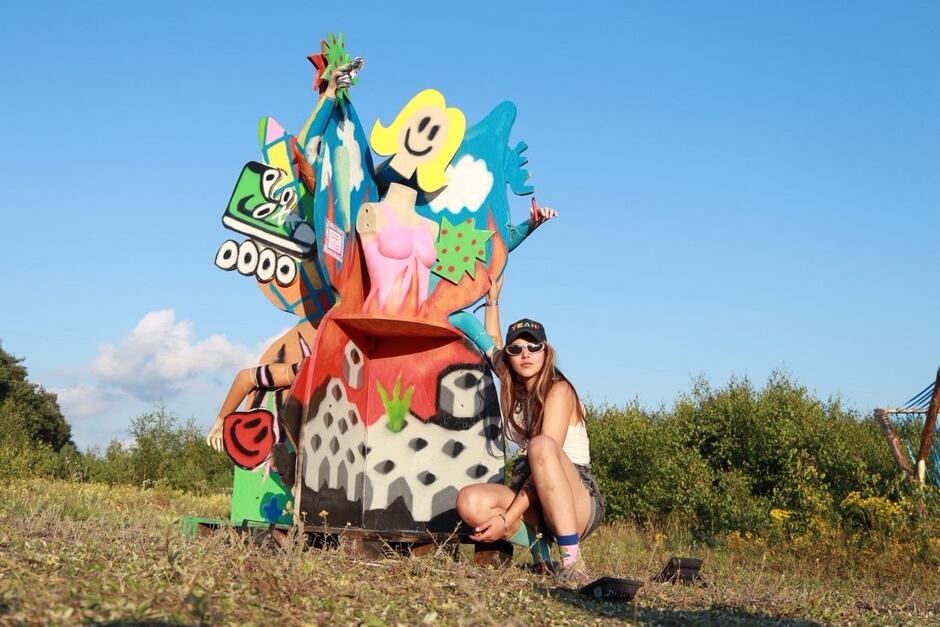 na zdjęciu kucająca kobieta przy kolorowej instalacji artystycznej, znajdują się na tle natury.