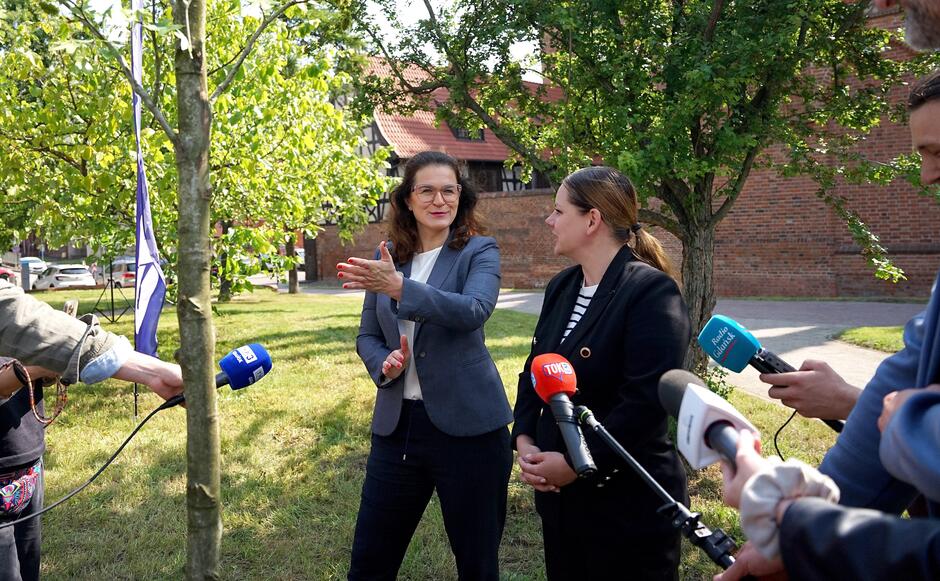 Dwie kobiety w średnim wieku stoją przy drzewku, które jest już posadzone z lewej strony i ma ponad 2 metry wysokości. Kobiety rozmawiają z dziennikarzami, widzimy mikrofony stacji radiowych i telewizyjnych