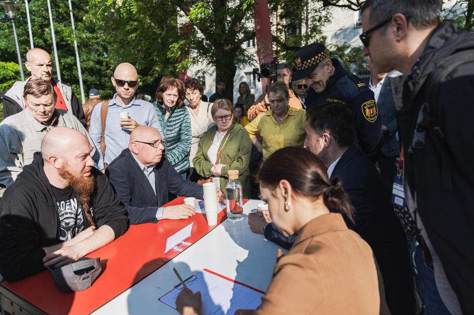 przy biało - czerwonym stole siedzi dwóch mężczyzn, naprzeciwko nich siedzą kobieta i mężczyzna, wokół nich stoi grupka kilkunastu osób i przysłuchuje się im