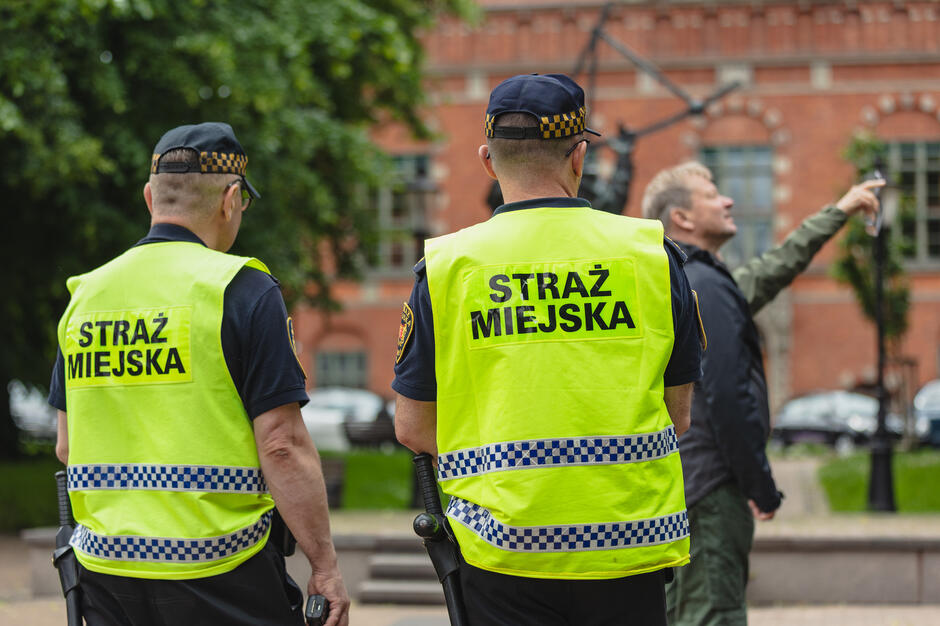 Dwóch strażników miejskich widzianych od tyłu stoi na ulicy miasta.