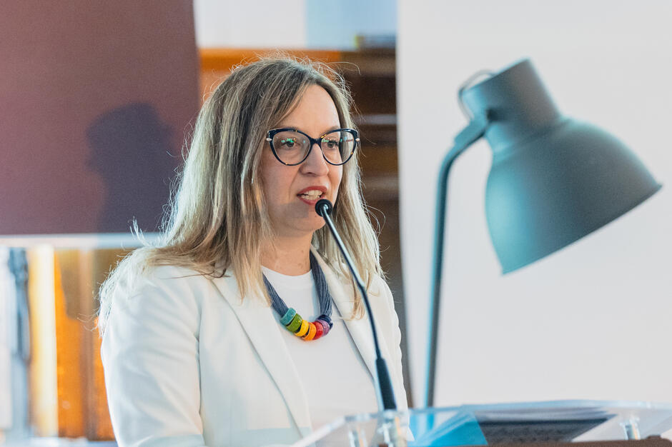 Kobieta w białym żakiecie przemawia do mikrofonu podczas oficjalnego wydarzenia. Ma długie, jasne włosy, okulary i kolorowy naszyjnik