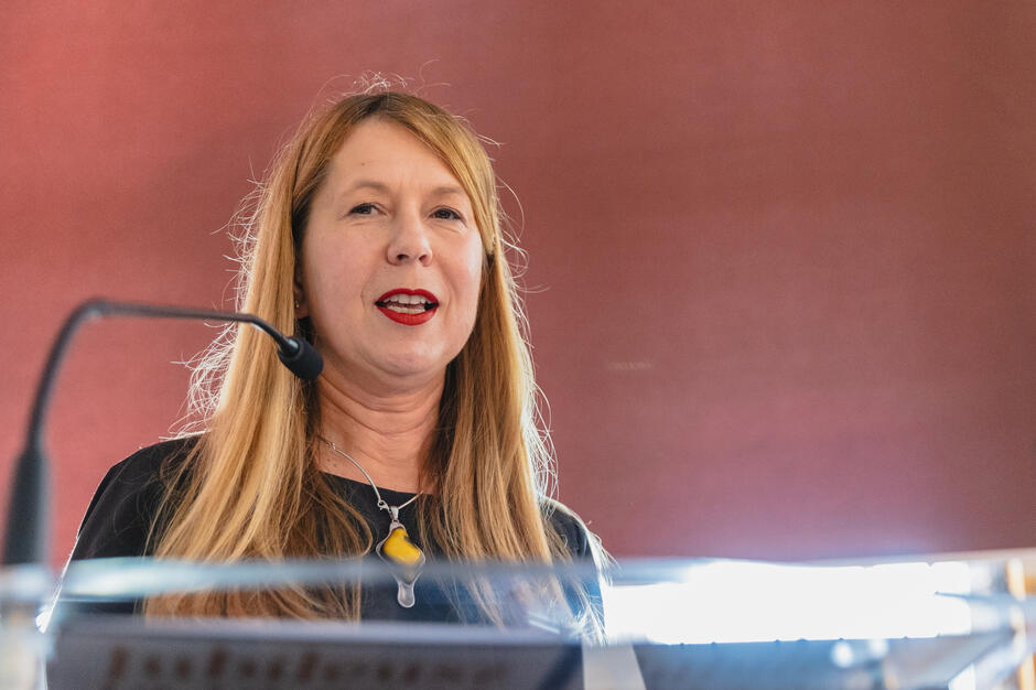 Kobieta w czarnej sukience z długimi, rudawymi włosami mówi do mikrofonu na tle czerwonej ściany. Ma wyrazisty makijaż i charakterystyczny naszyjnik z żółtym akcentem