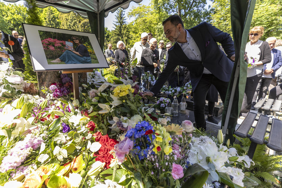 Dużo kwiatów na grobie, mężczyzna w średni wieku kładzie kolejną wiązankę