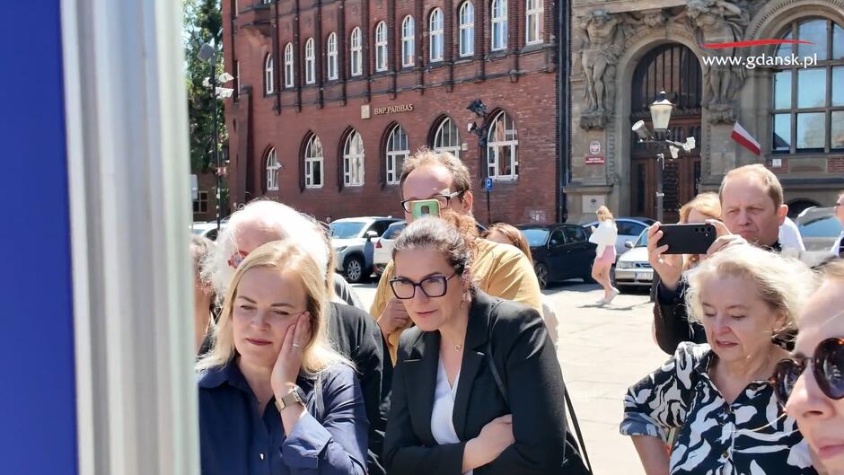 Grupa osób przygląda się zdjęciom na wystawie, na pierwszym palnie trzy osoby