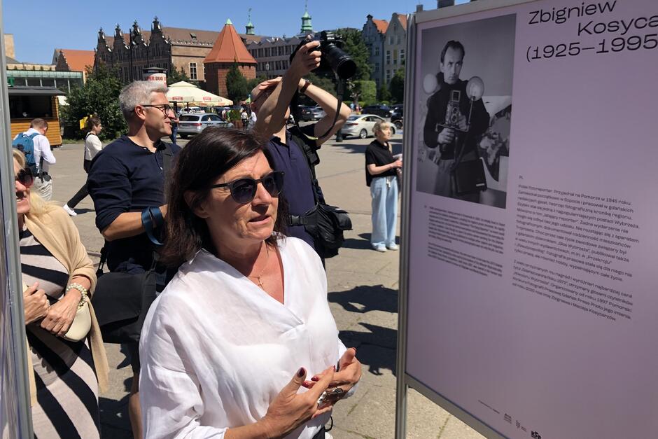 Kobieta w okularach przeciwsłonecznych opowiada gościom o zdjęciach wybranych na wystawę