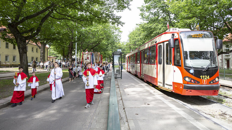 grupa ludzi w kolorowych strojach idzie po ulicy niosąc udekorowane feretrony. Obok, na przystanku widoczny tramwaj czekający na pasażerów